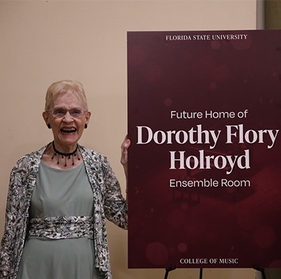 Dorothy Flory Holroyd stands smiling next to a sign about the dedication ceremony on her behalf